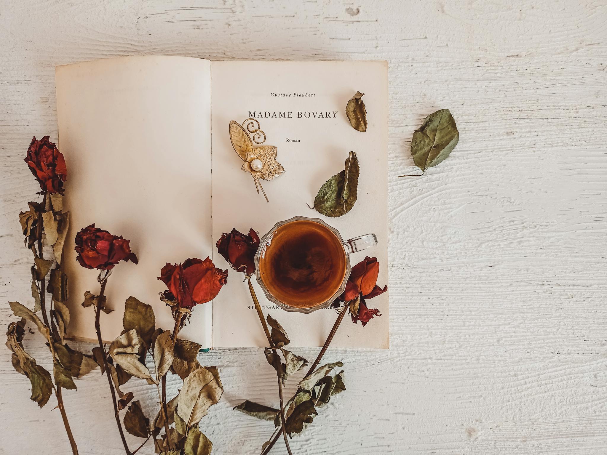 Aesthetic flat lay featuring Madame Bovary, a brooch, tea, and dried roses on a wooden surface.