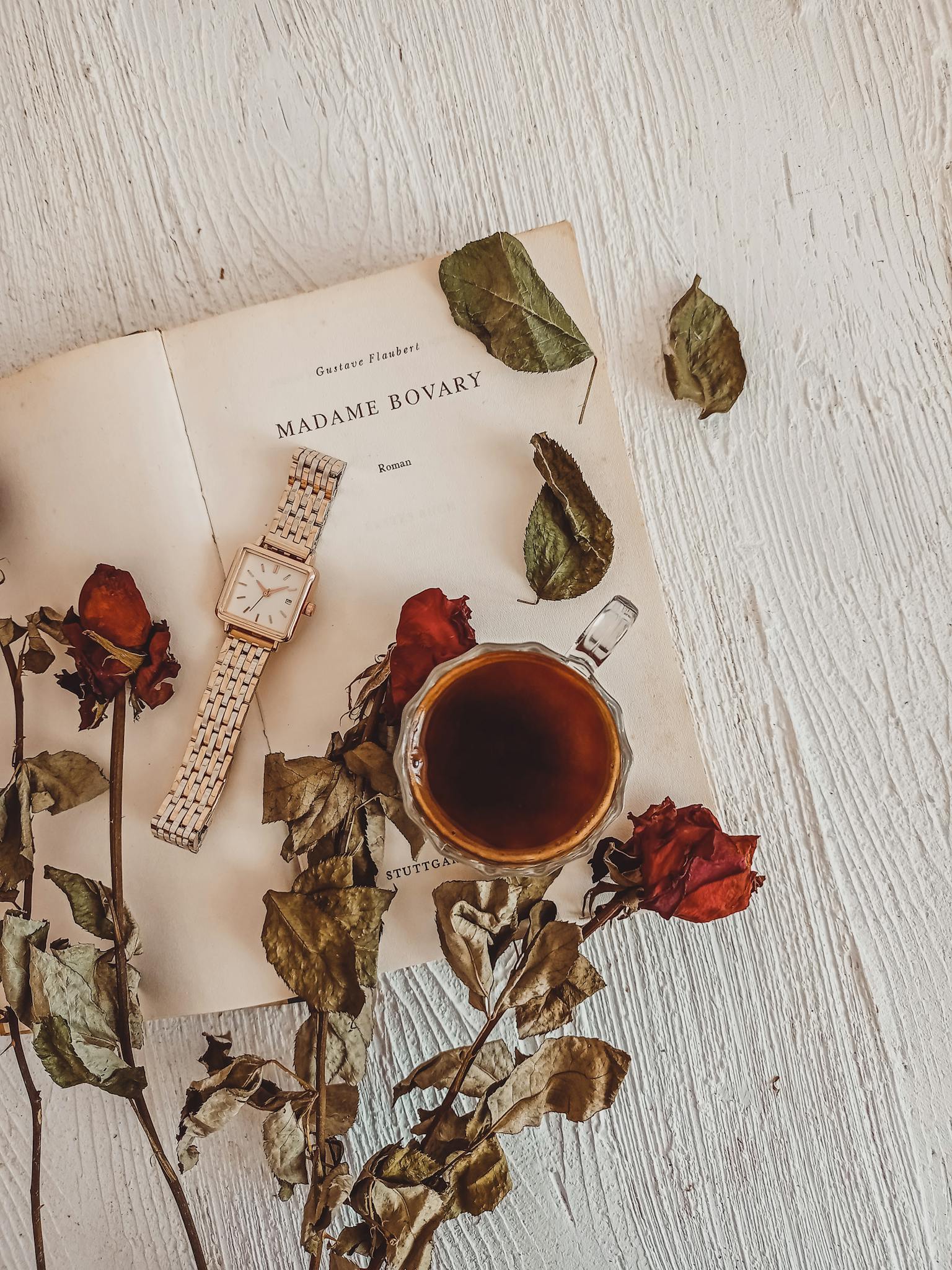 Charming flat lay featuring an open book, wristwatch, teacup, and dried roses on wooden surface.