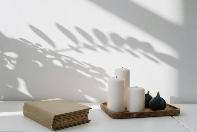 Elegant still life featuring candles, a book, and leaf shadows on a white tabletop.